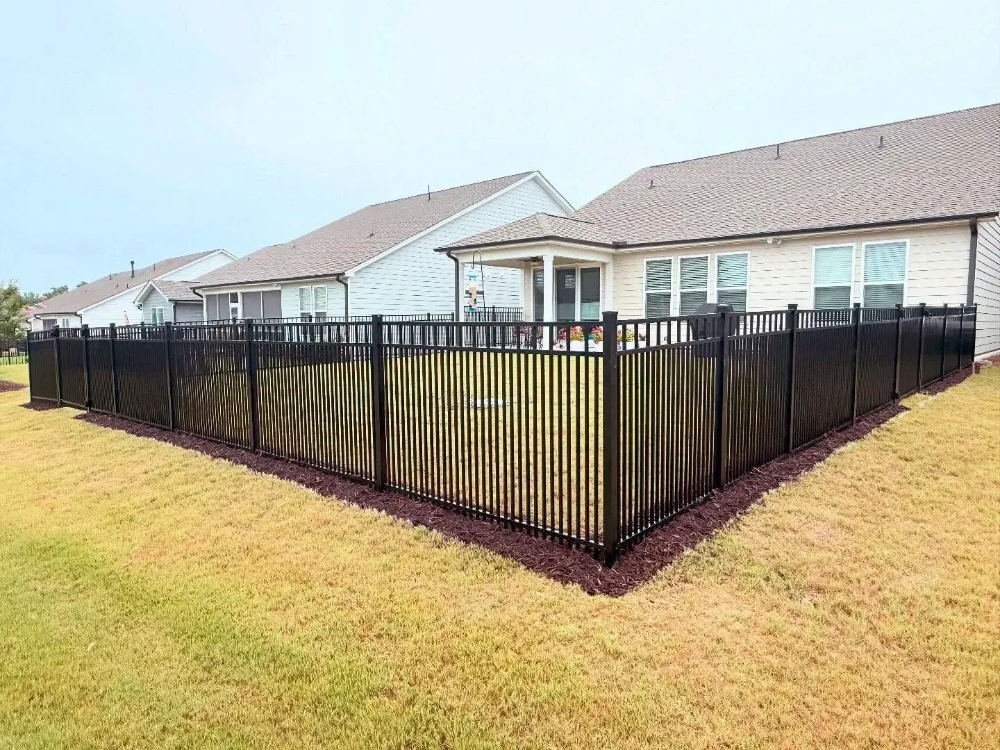 Black metal fence surrounds a grassy yard in front of multiple houses under a cloudy sky.