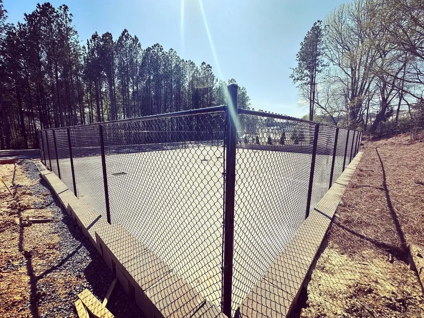 Black net fence enclosure on a concrete base, in a wooded area on a sunny day.