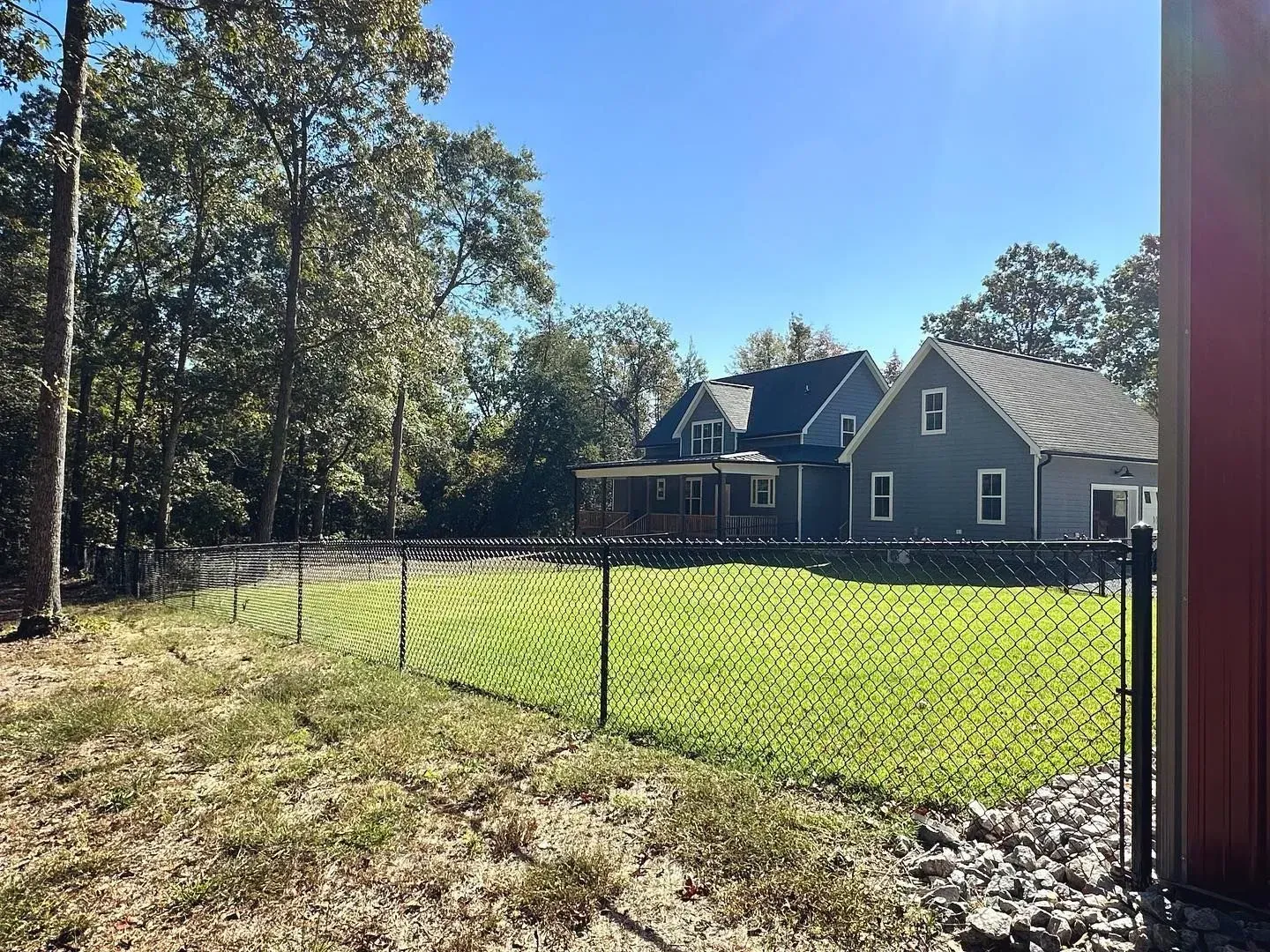 Chain-link fence in front of a green lawn and two-story houses under a sunny, blue sky.