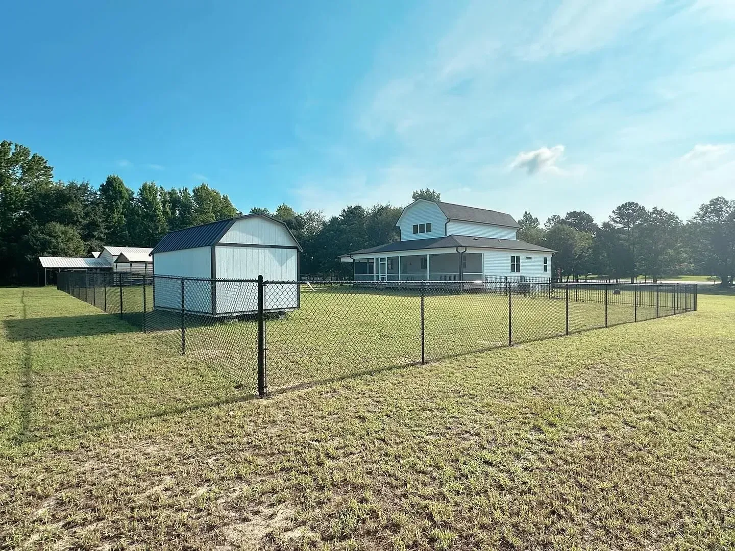 A fenced backyard with a house, shed, and trees under a blue sky.