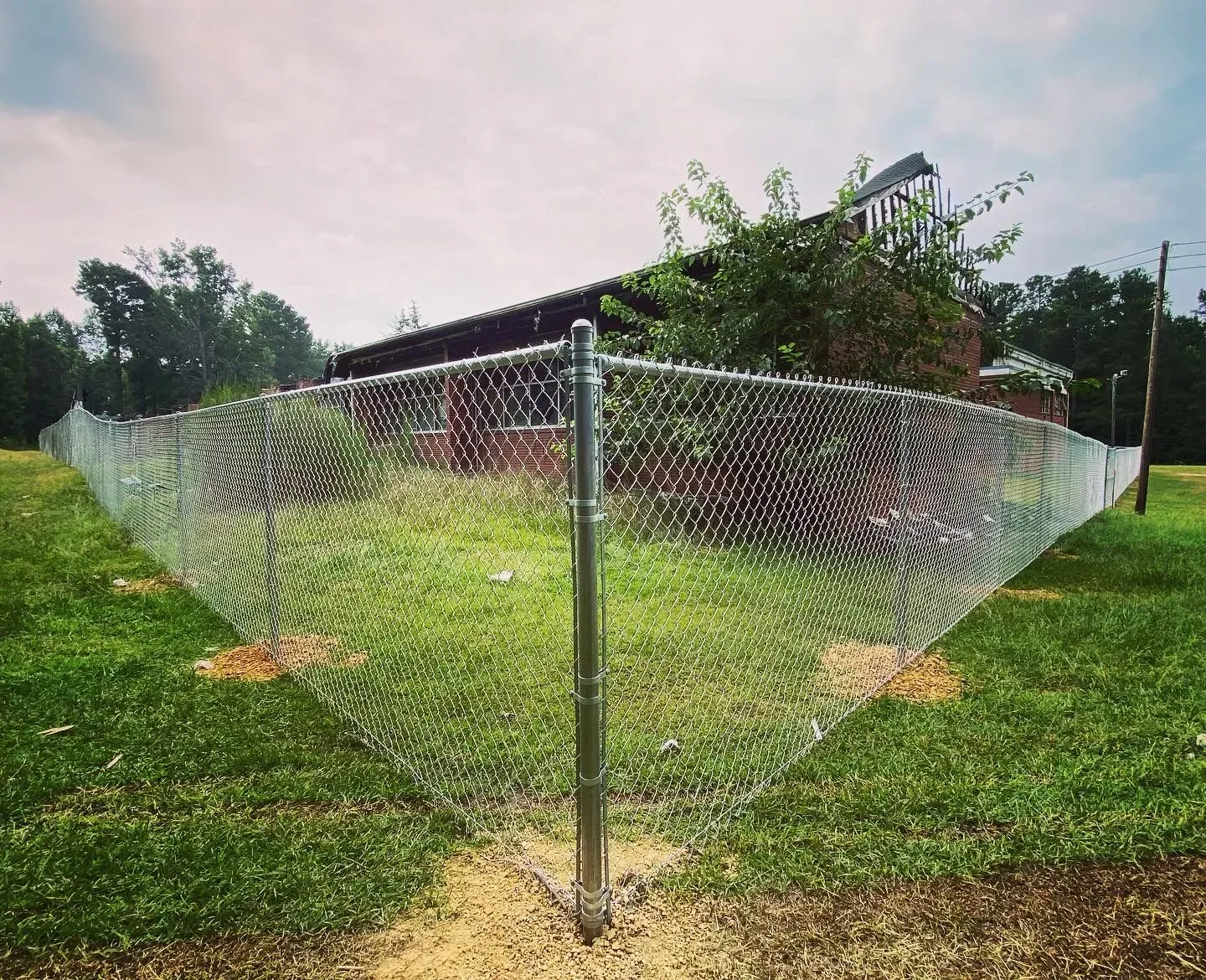 Chain-link fence in front of a brick building on a grassy lawn under a cloudy sky.
