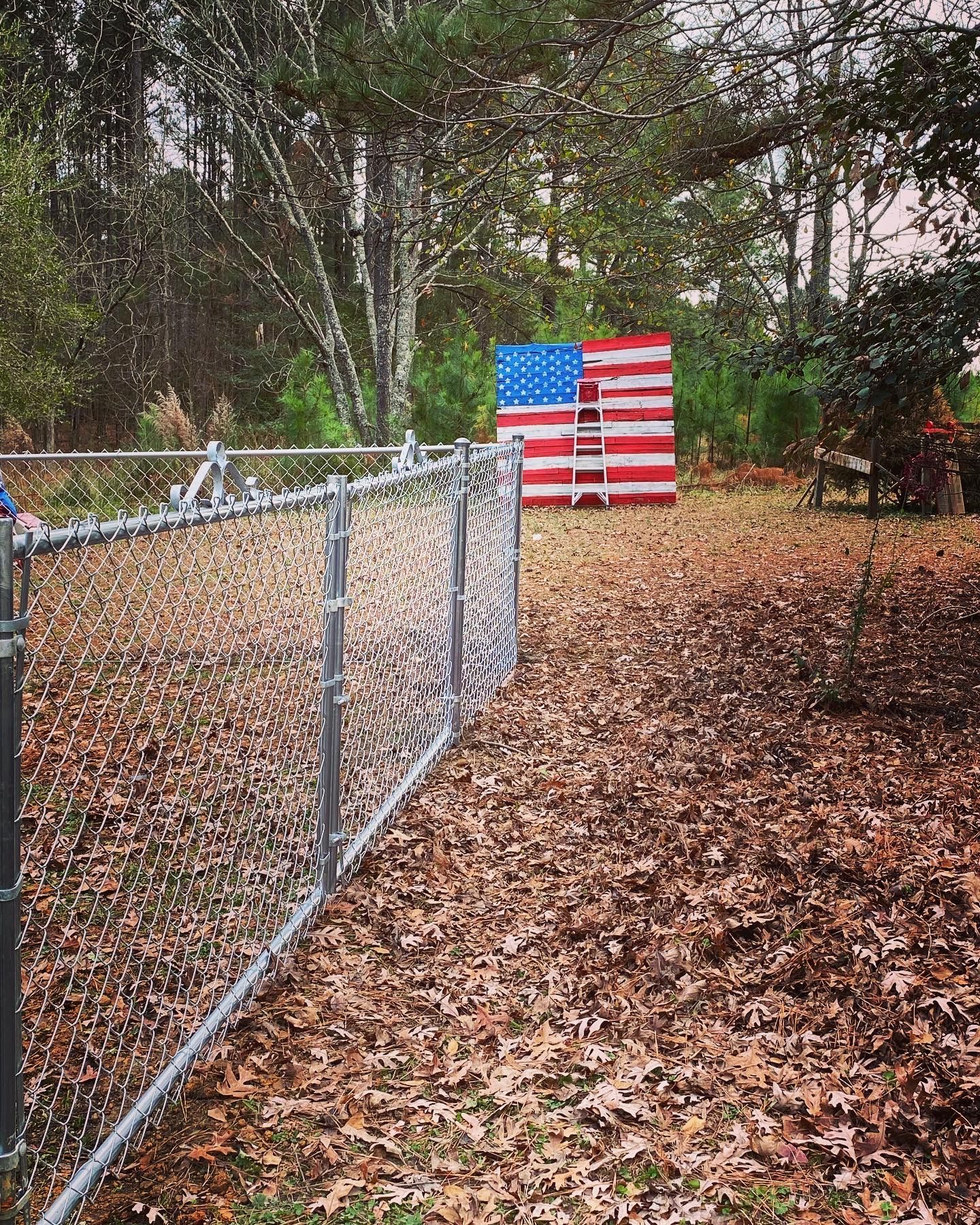 American flag painted on a wooden panel, behind a chain-link fence, in a wooded area.