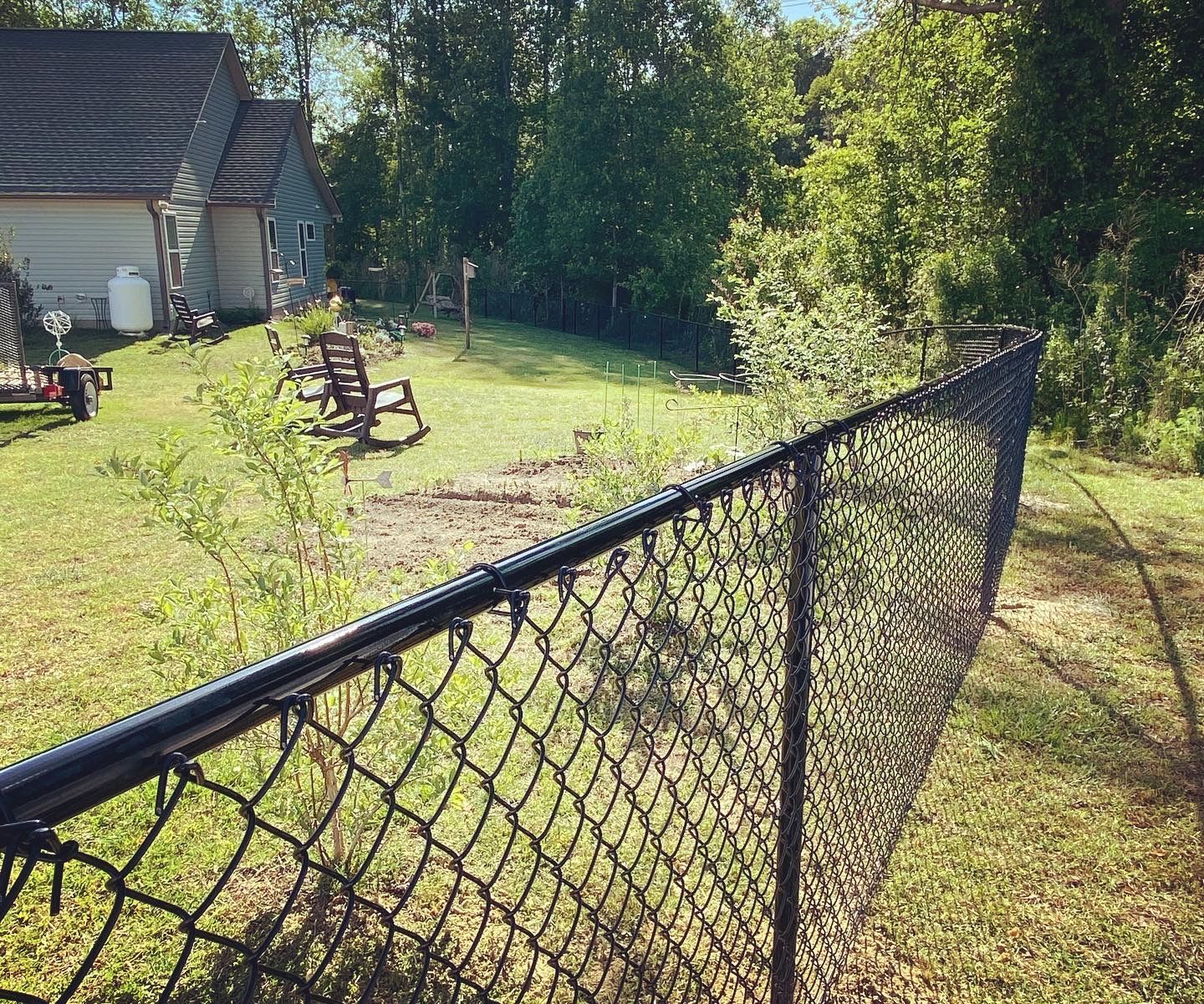 Black chain-link fence bordering a grassy backyard with a house, trees, and two chairs.