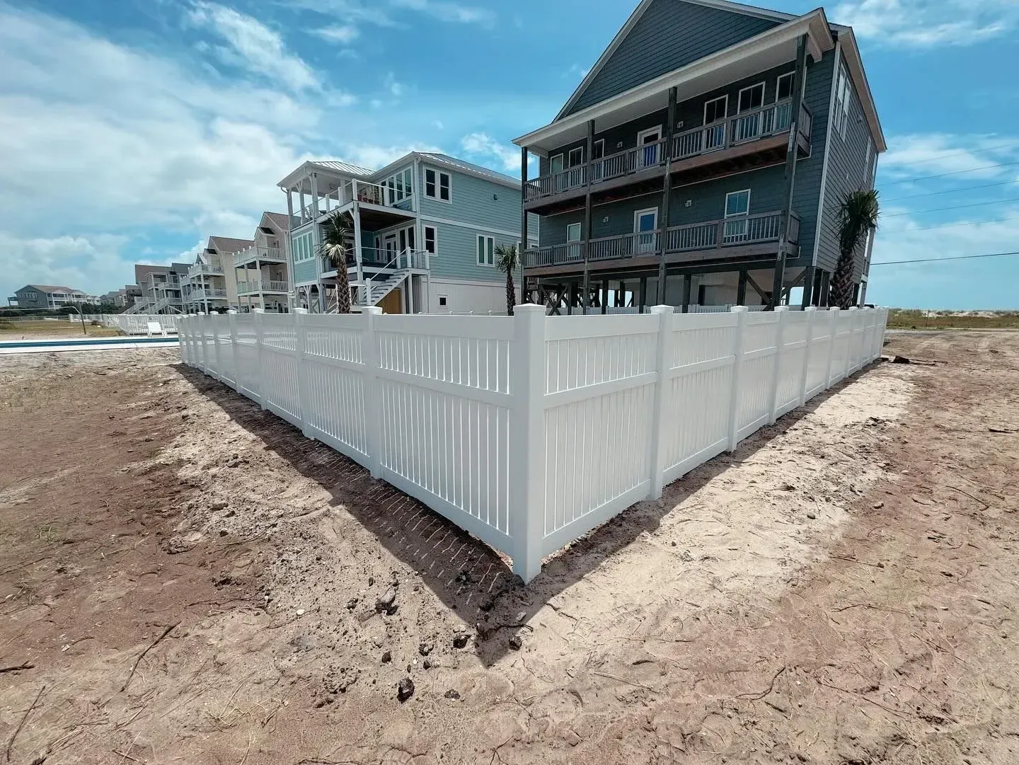 White fence encloses beach houses on a sandy beach under a bright blue sky.