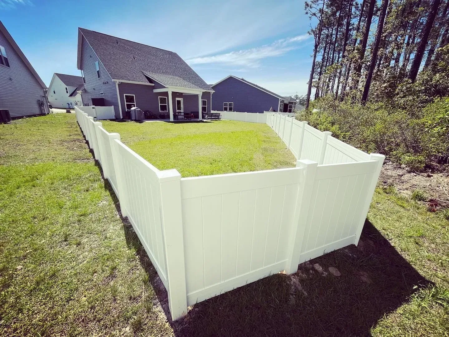 White vinyl fence encloses a backyard with a house, green grass, and trees under a blue sky.