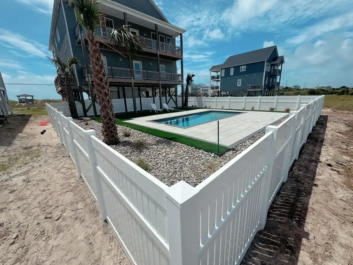 Beach house with a white fence around a pool on a sandy lot.