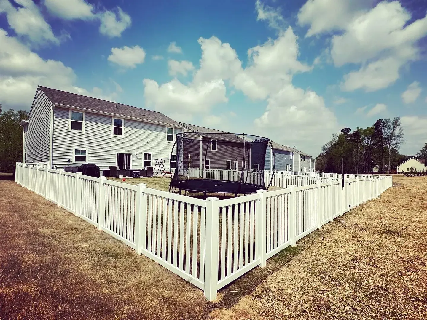 White picket fence encloses a backyard with a two-story house, under a blue sky with clouds.