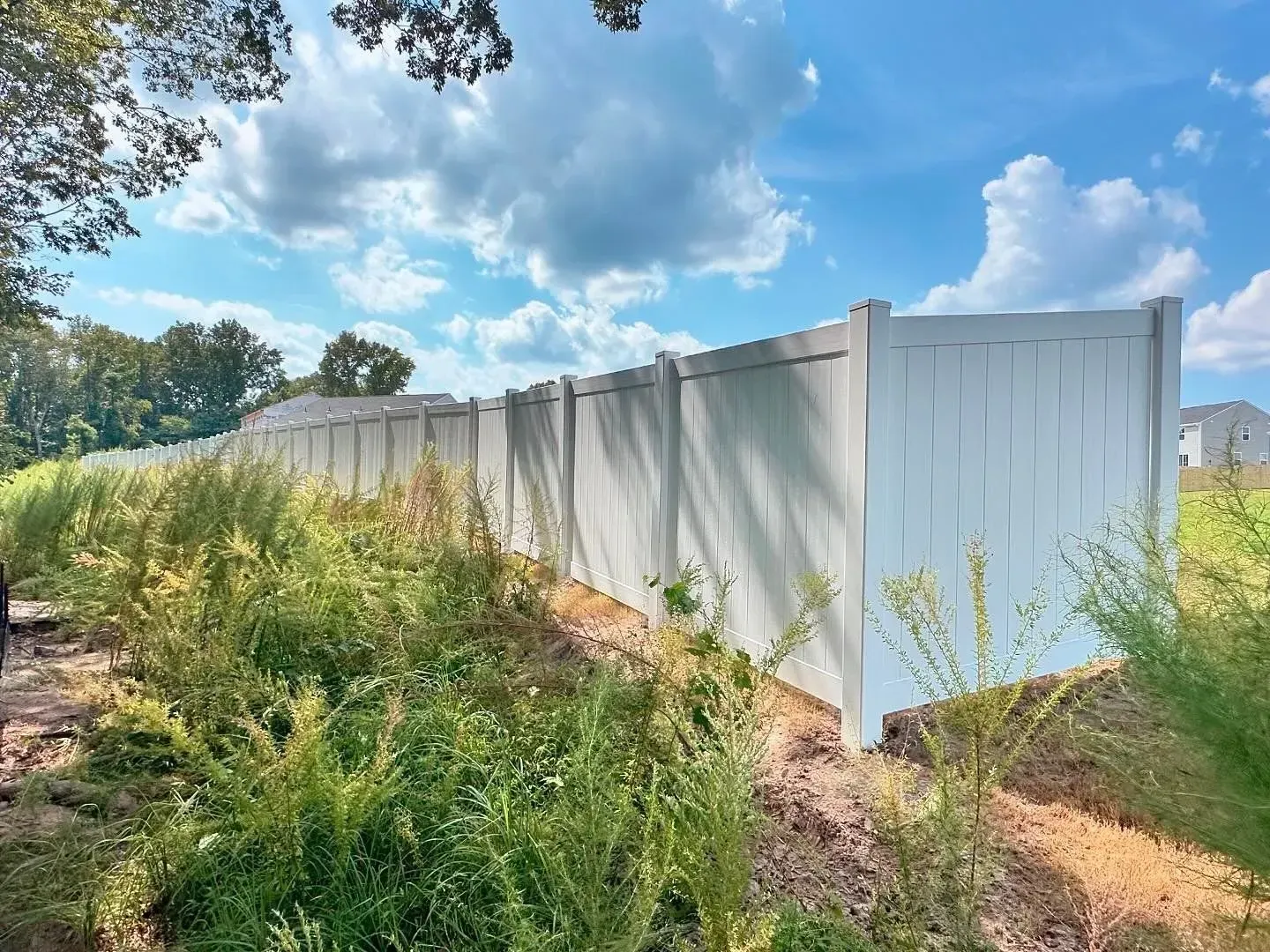 White vinyl fence next to overgrown plants under a partly cloudy sky.