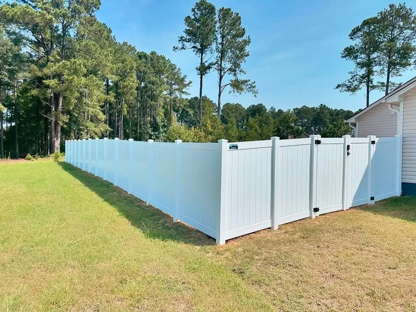 White vinyl fence surrounds a grassy yard on a sunny day, with trees in the background.