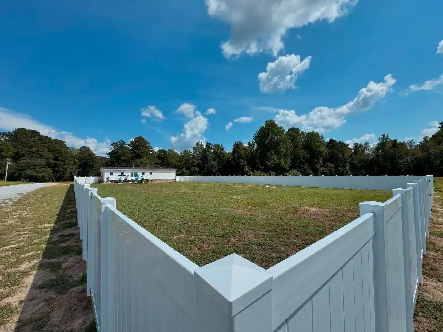 White fenced-in yard with green grass, small building, and blue sky with clouds.