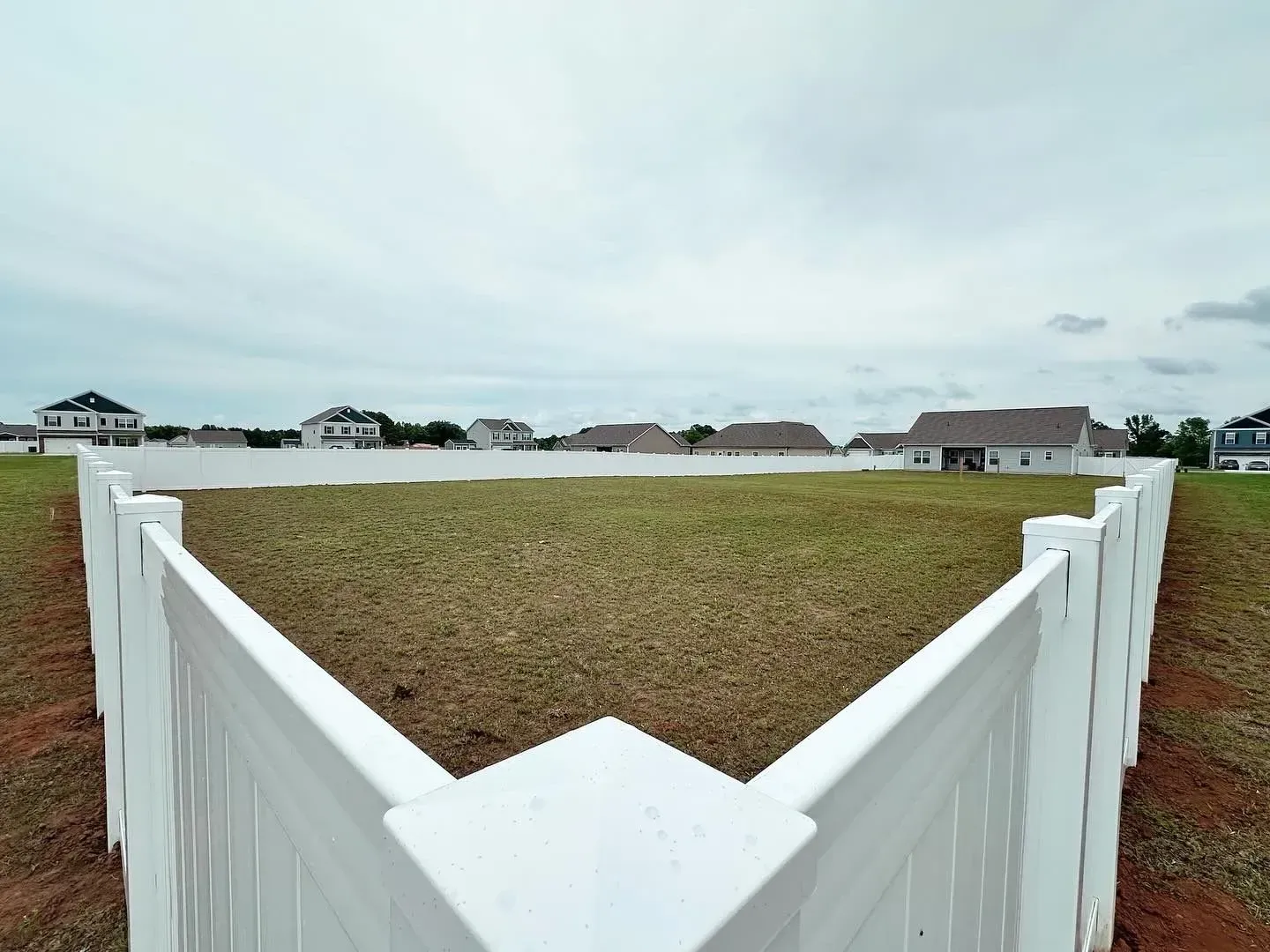 White picket fence surrounding a large, empty lot with grass, houses in the distance, overcast sky.