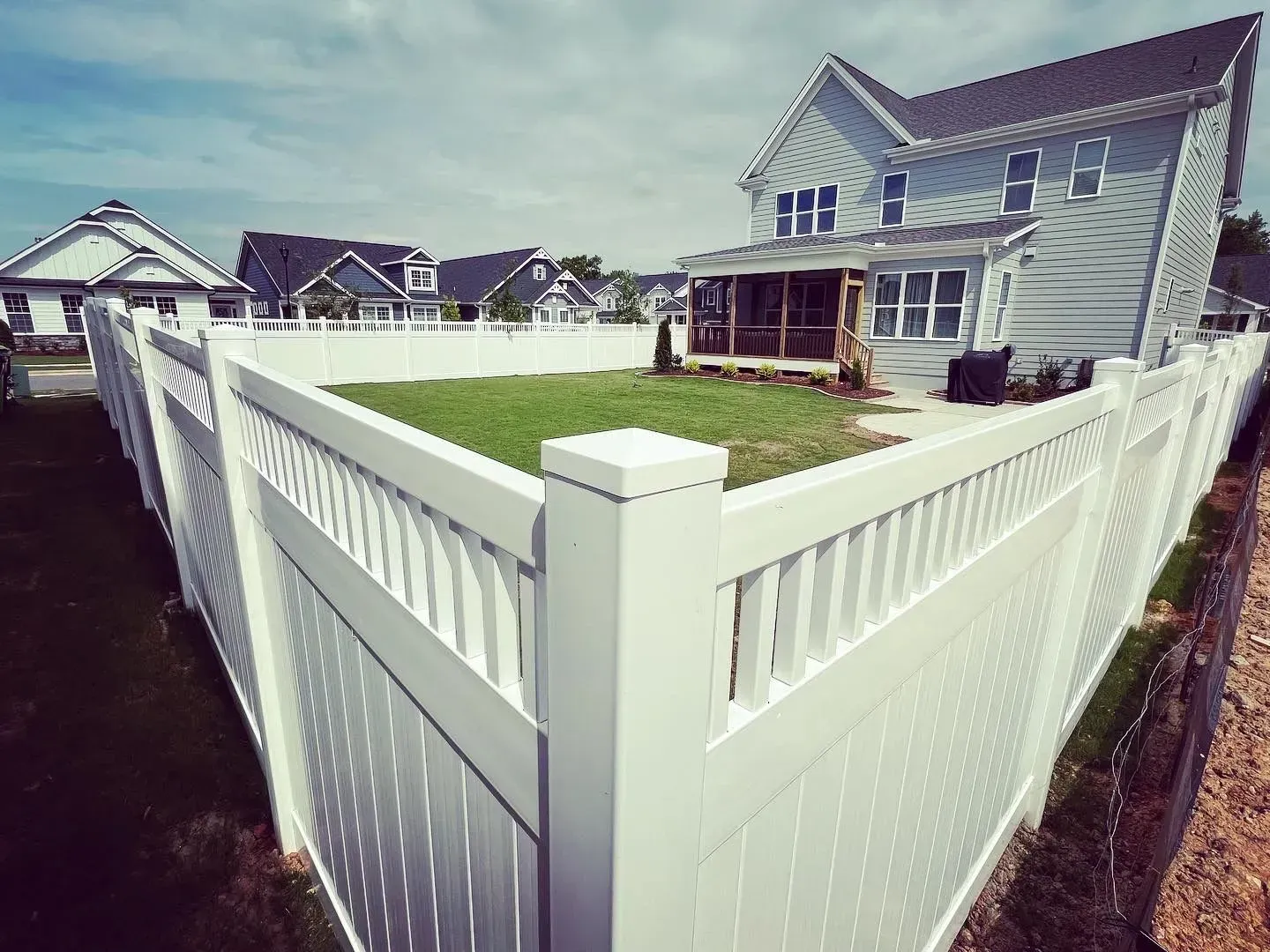 White vinyl fence surrounding a grassy backyard with a house, neighborhood in the background, blue sky.