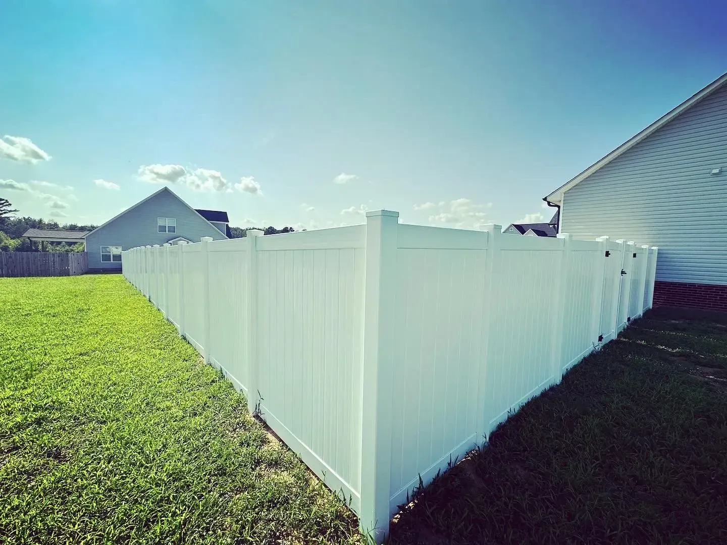 White vinyl fence encloses a green lawn, adjacent to a house, under a blue sky.