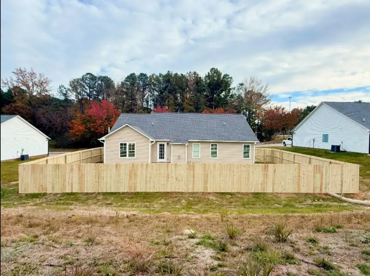 Tan house with gray roof enclosed by a wooden fence, set in a field.