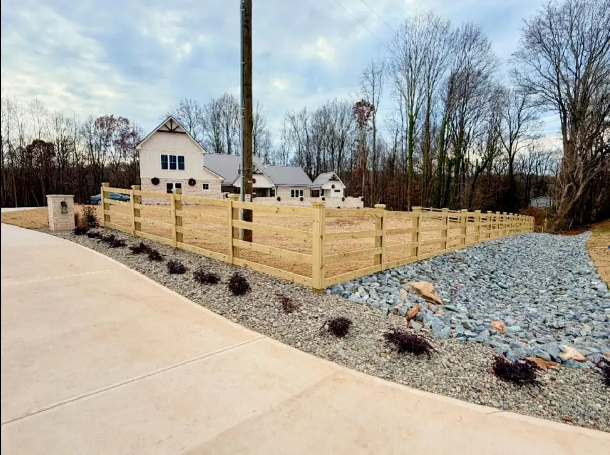 Wooden fence surrounding a house with landscaping, a gravel driveway, and cloudy sky.