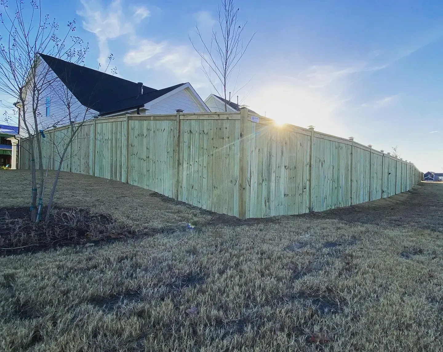 Wooden fence bordering a grassy yard with a house and bright sun in the background.