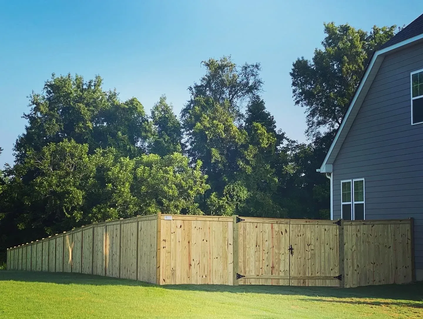 Wooden fence surrounding a grassy yard next to a gray house, trees in background.