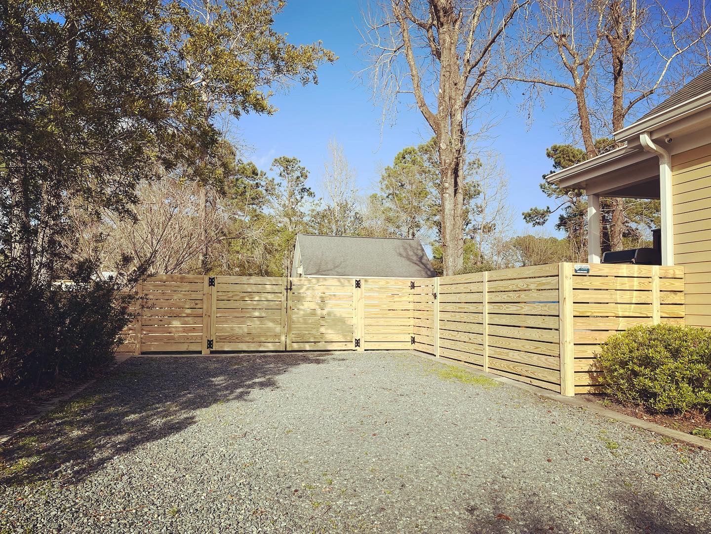 Gravel driveway framed by wooden slatted fence, trees, and a house under a clear blue sky.