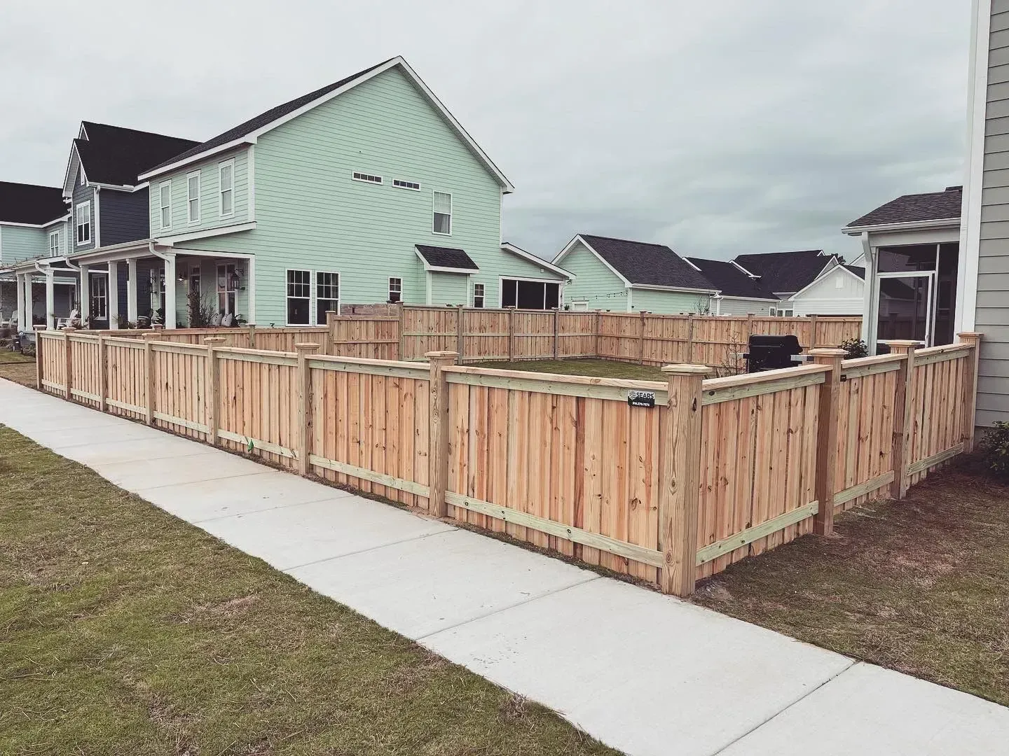 Wooden fence surrounding a grassy yard next to a sidewalk and houses.