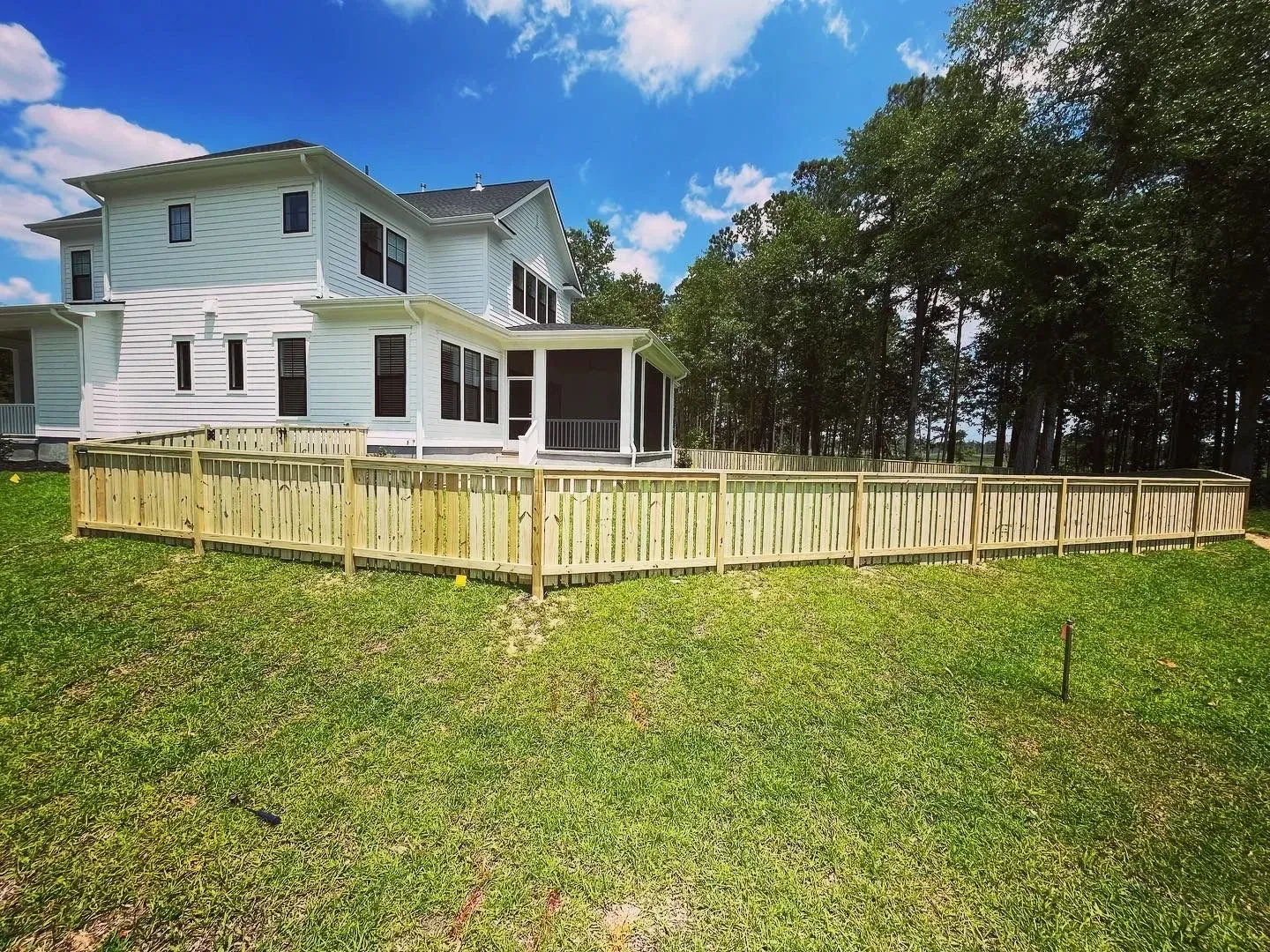 Wooden fence surrounds a two-story white house with a screened porch on a grassy lawn under a blue sky.
