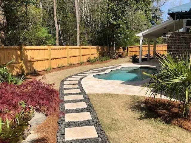 Stone path leads past landscaping to a pool in a backyard with a wooden fence.