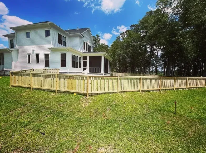 White house with a wooden fence in a grassy yard under a blue sky with trees.