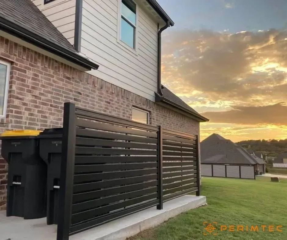 Black slatted privacy fence beside a brick house, concealing trash cans, with sunset background.