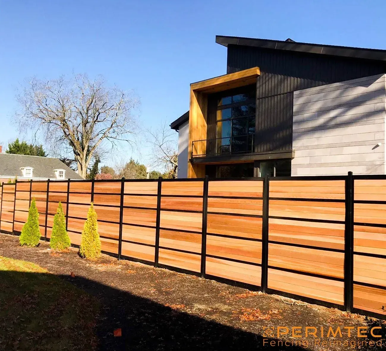 Wooden fence with black posts, in front of a modern house, with small trees and blue sky.