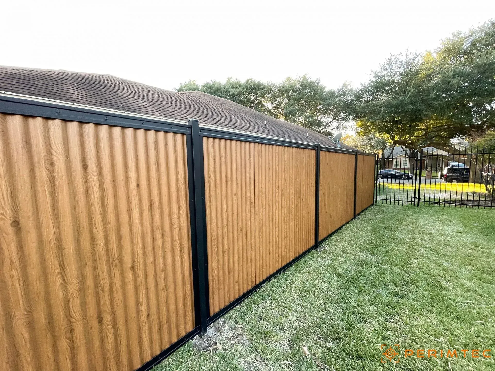 A wooden-look fence with black framing encloses a backyard; green grass and trees are in the background.