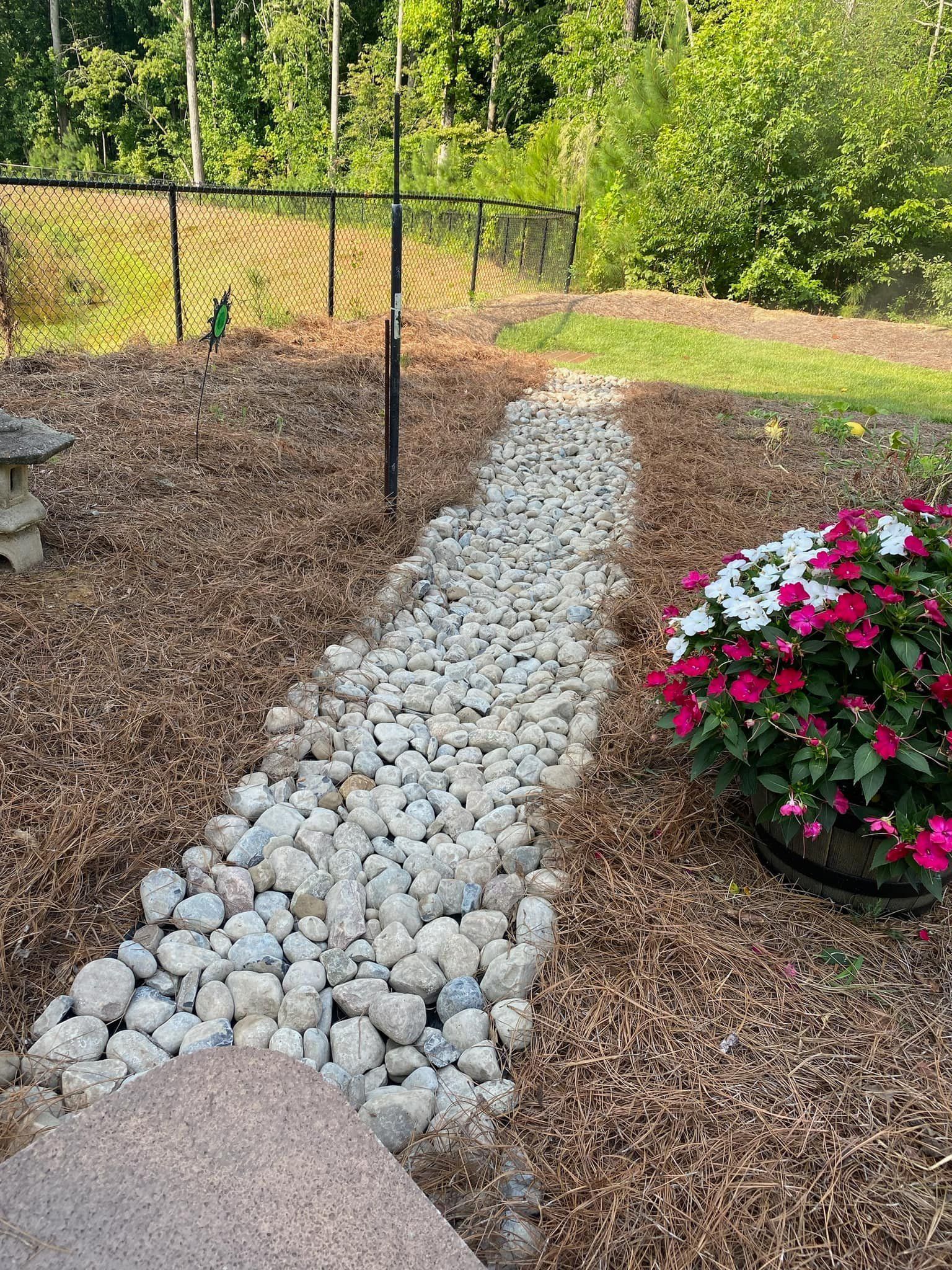 A rock-lined pathway through wood chips, with a potted plant on the right and a fence in the background.