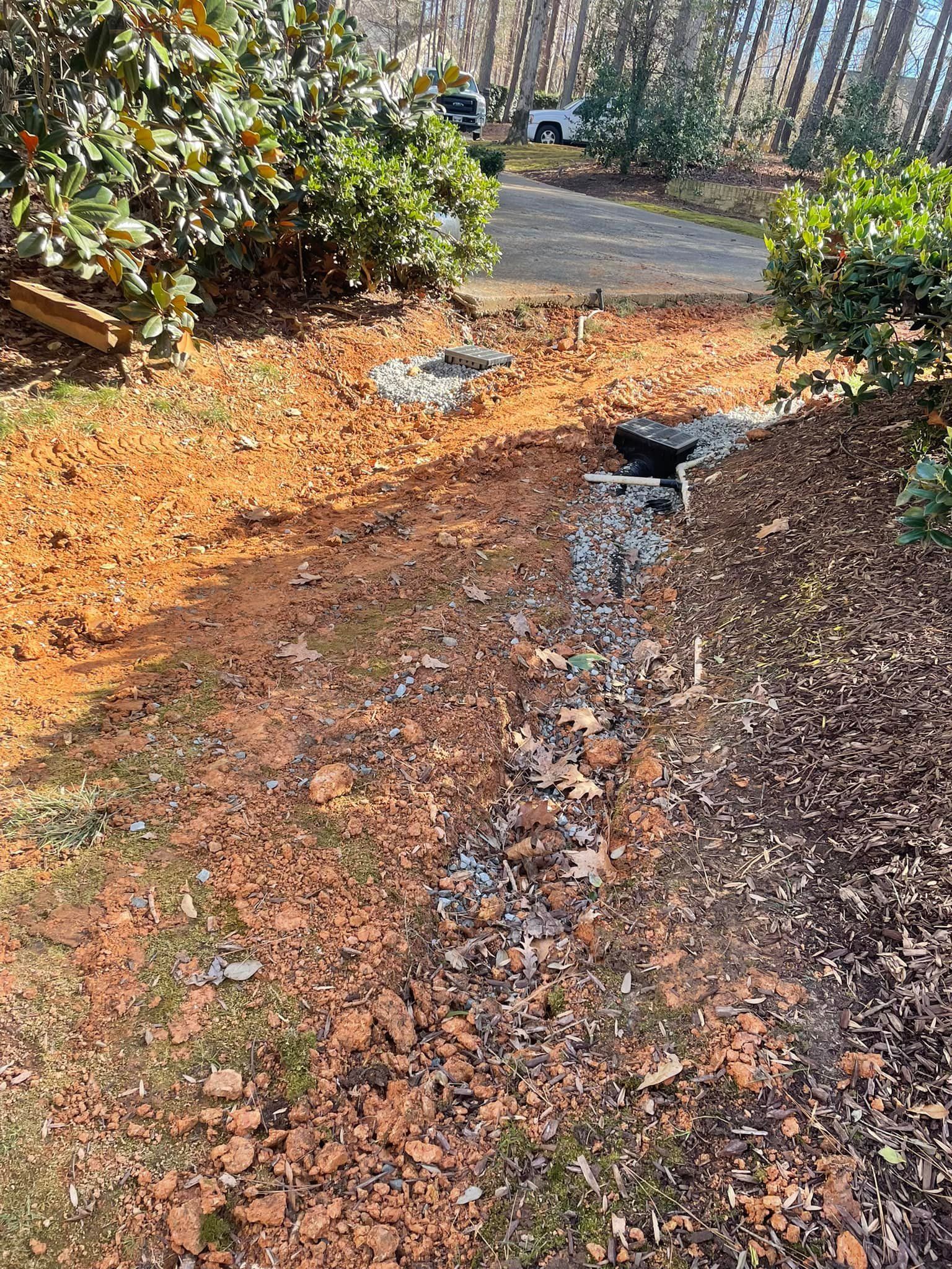 Drainage ditch with gravel and leaves, leading to a black drain, near bushes and a driveway.