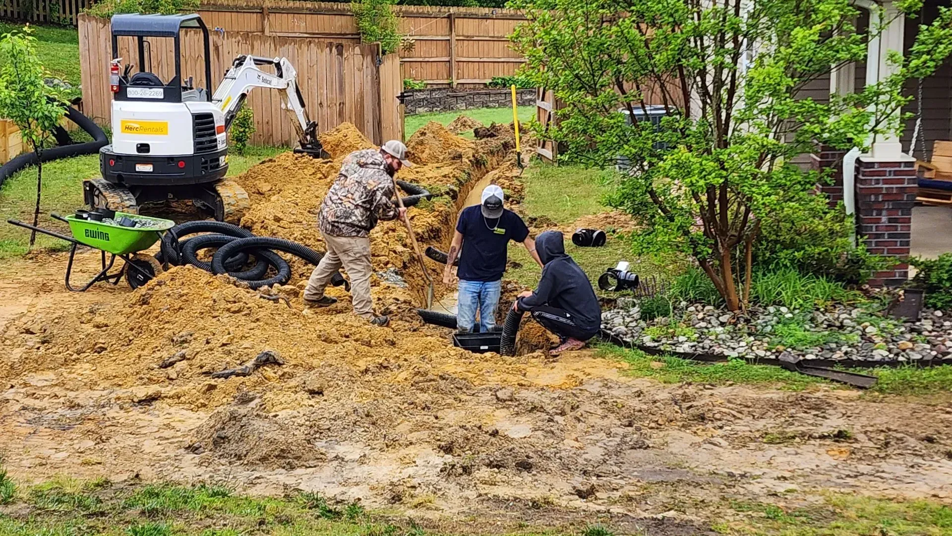 Construction workers installing drainage pipes near a house, with an excavator and equipment in the mud.