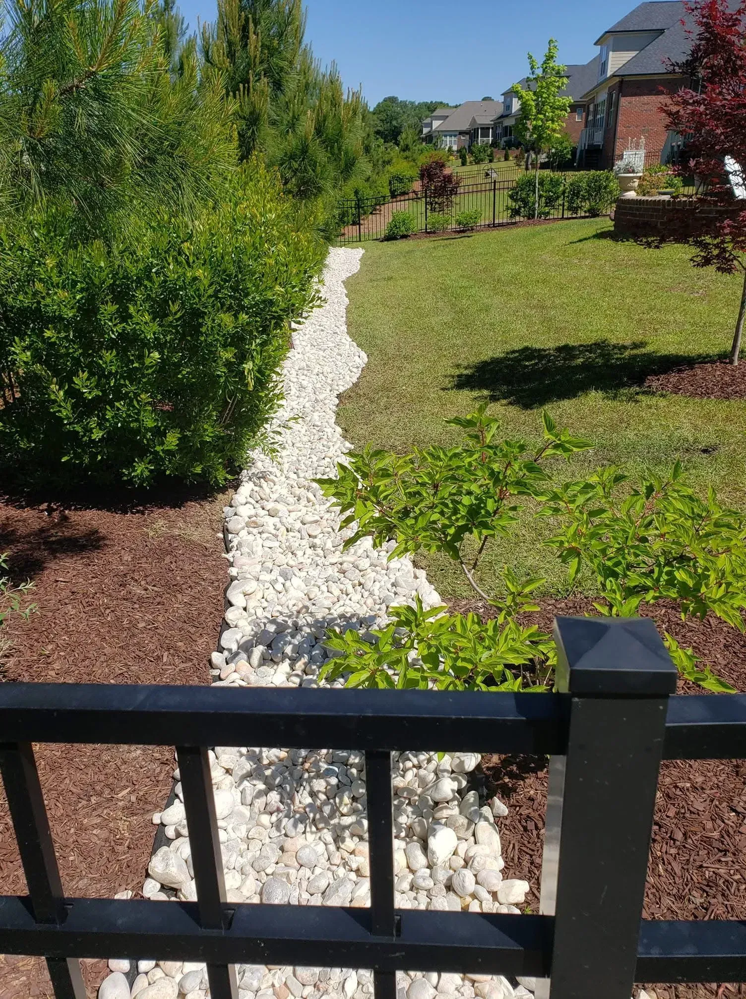A backyard drainage channel lined with rocks, bordered by shrubs, mulch, and a black fence.
