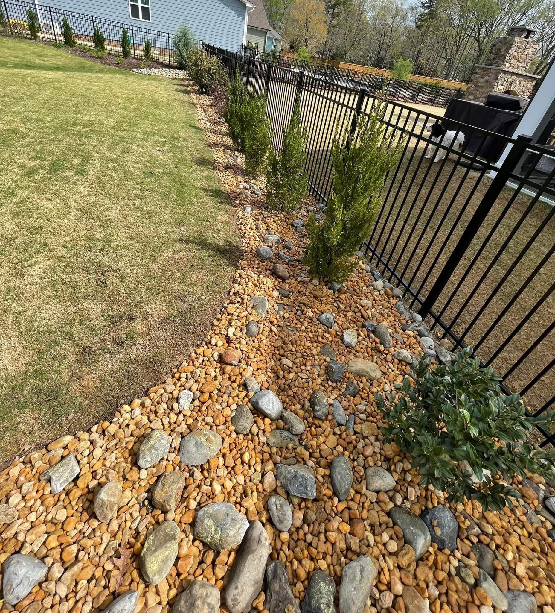 Yard with black fence, brown gravel and rocks, green grass, and small shrubs.