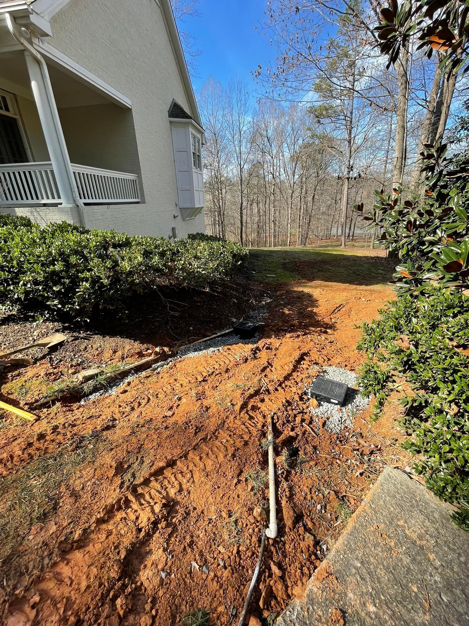 Excavated trench next to a house with exposed pipe and red dirt. Overcast sky with trees in the background.