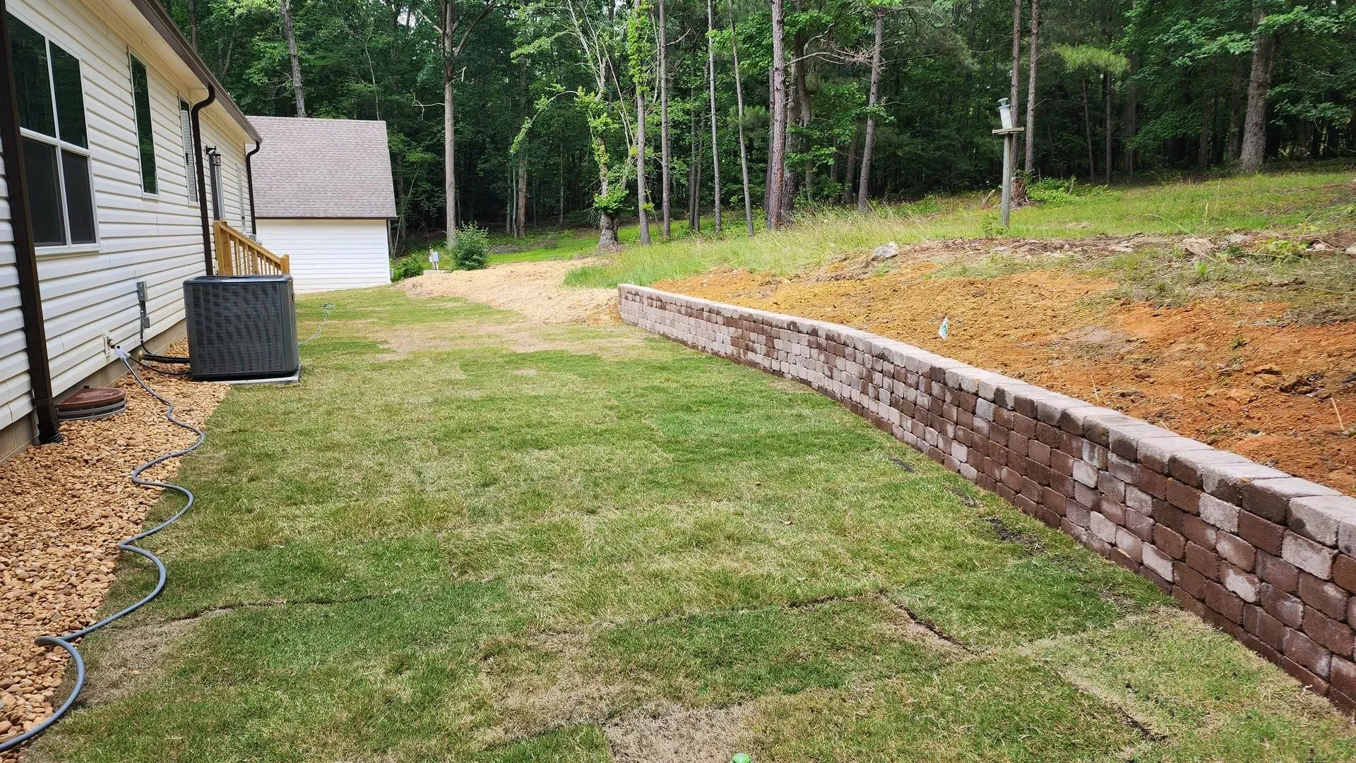 A brick retaining wall borders a backyard with grass. Trees in the background. White house on the left.
