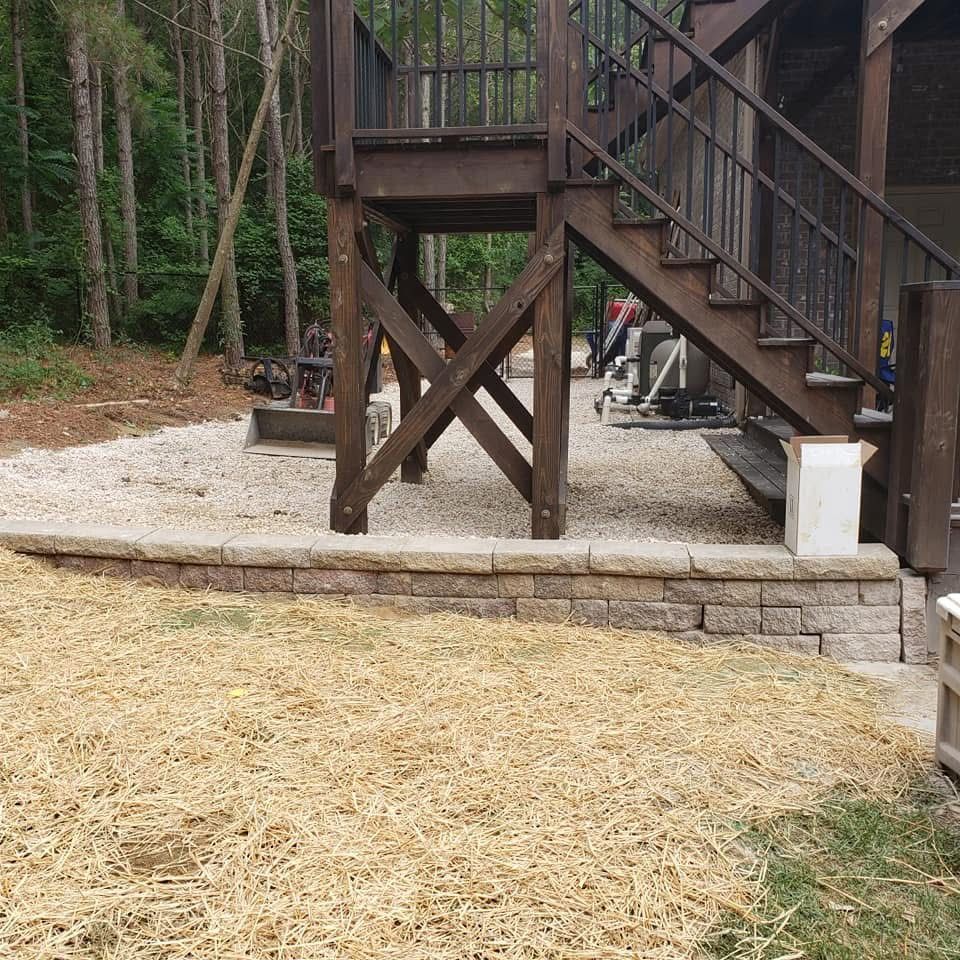 Wooden stairs leading up to a deck supported by beams, resting on a retaining wall and gravel.