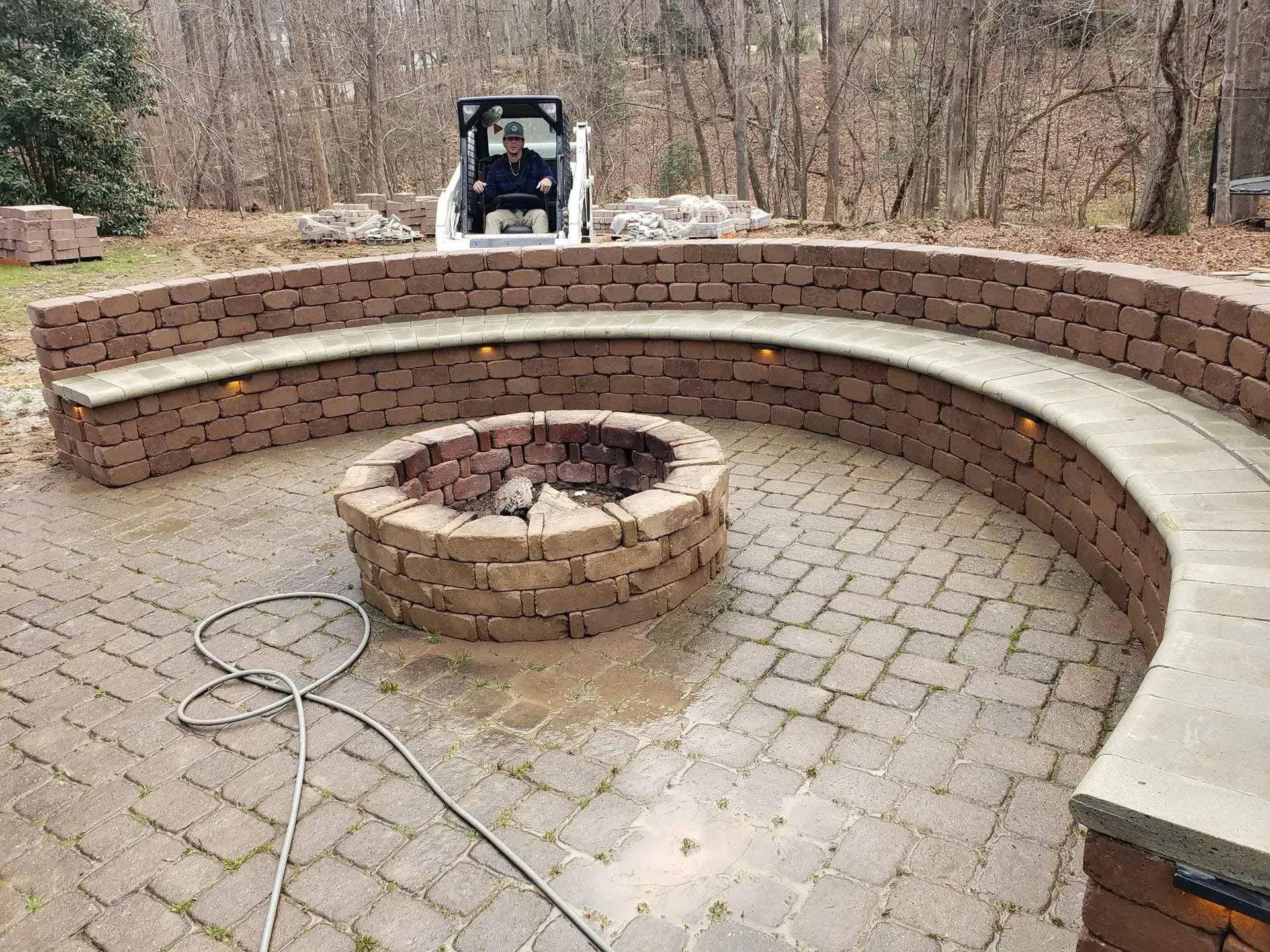 Brick fire pit with curved brick seating on a brick patio. A man in a skid steer is in the background.