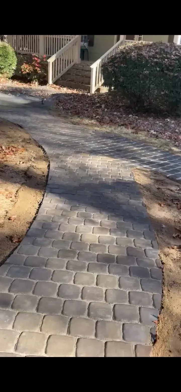 Brick paved pathway curves toward a house with a staircase, framed by shrubs and trees.