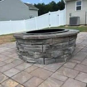 Stone fire pit on a patio, with a white fence and house in the background.