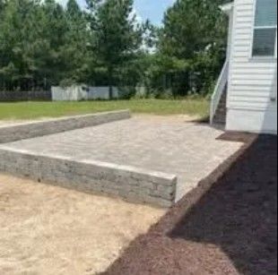 Stone patio with retaining walls next to a house with stairs, set in a yard with trees and mulch.