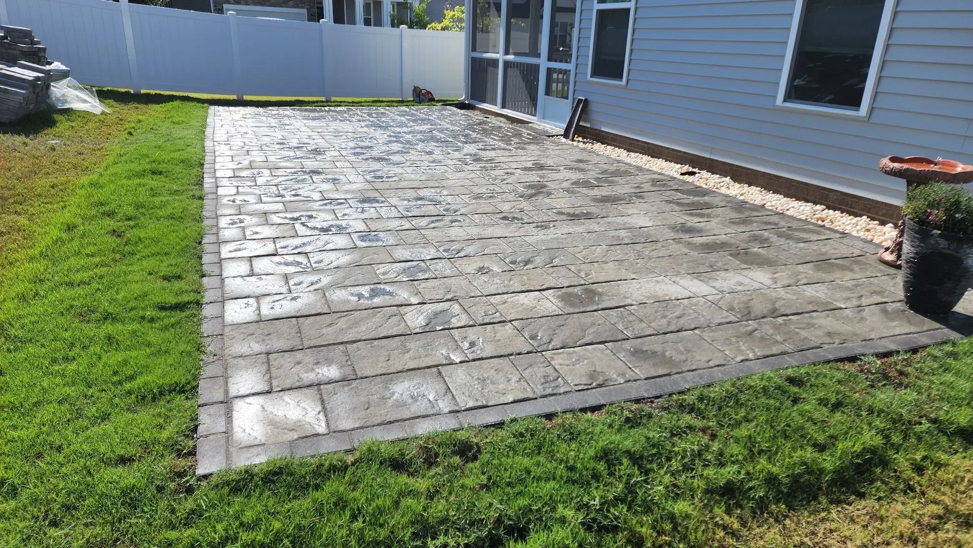 Paver patio bordered by grass next to a light blue house and a white fence.
