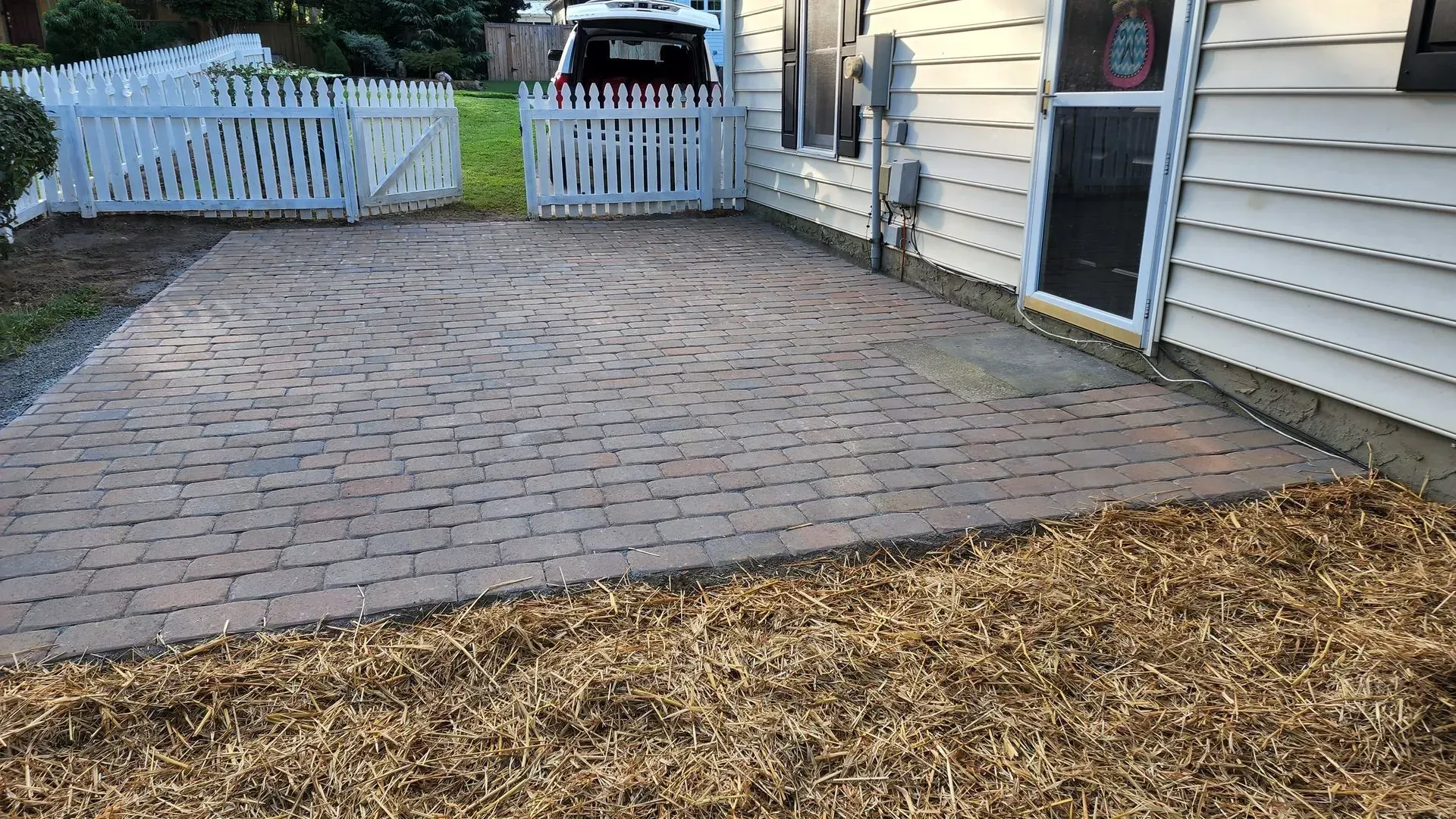 Brick patio with curved pattern, adjacent to a house and white picket fence.