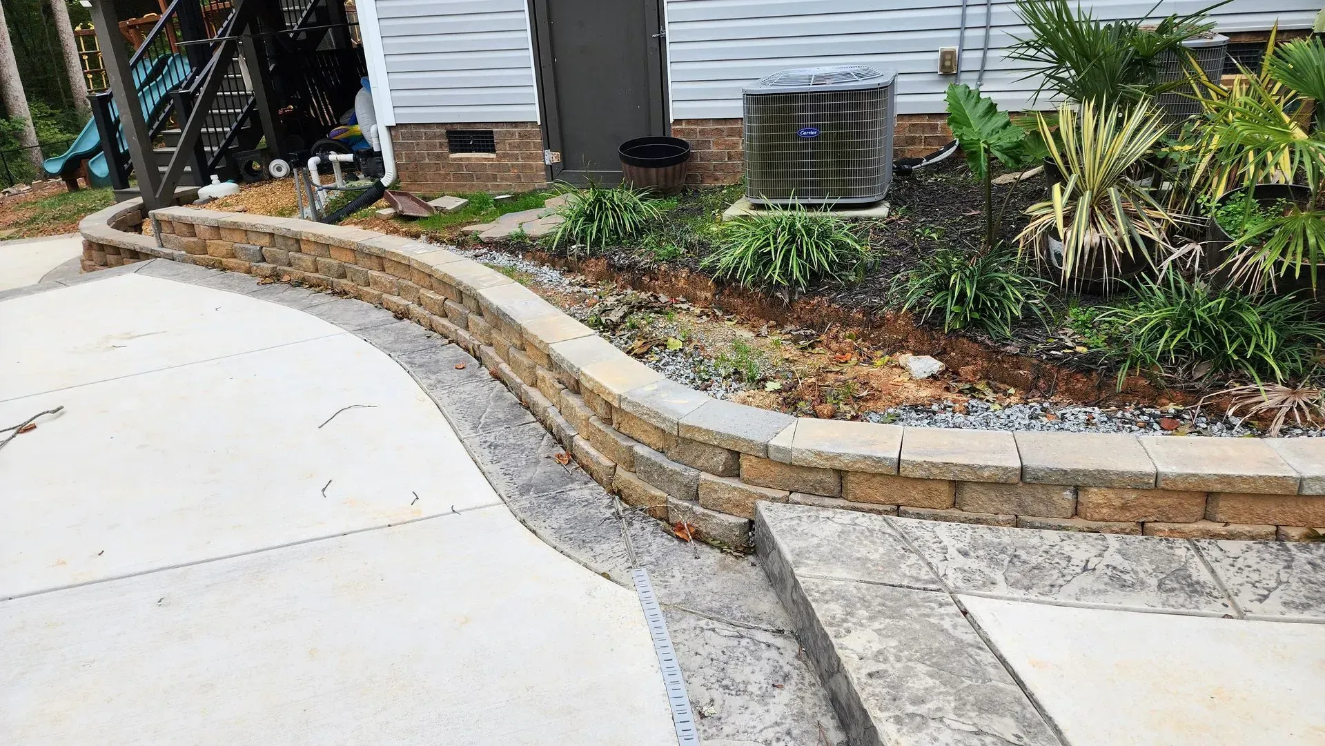 Curved retaining wall made of tan bricks next to a concrete driveway and garden bed with plants.