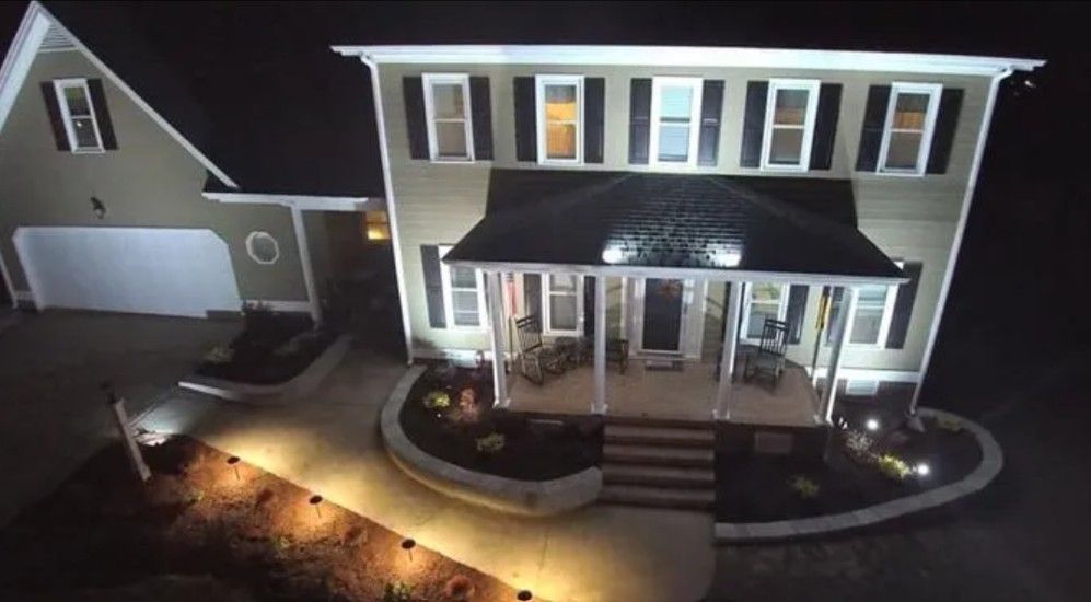 Night view of a two-story house with illuminated porch, walkway, and landscaping. Dark sky.