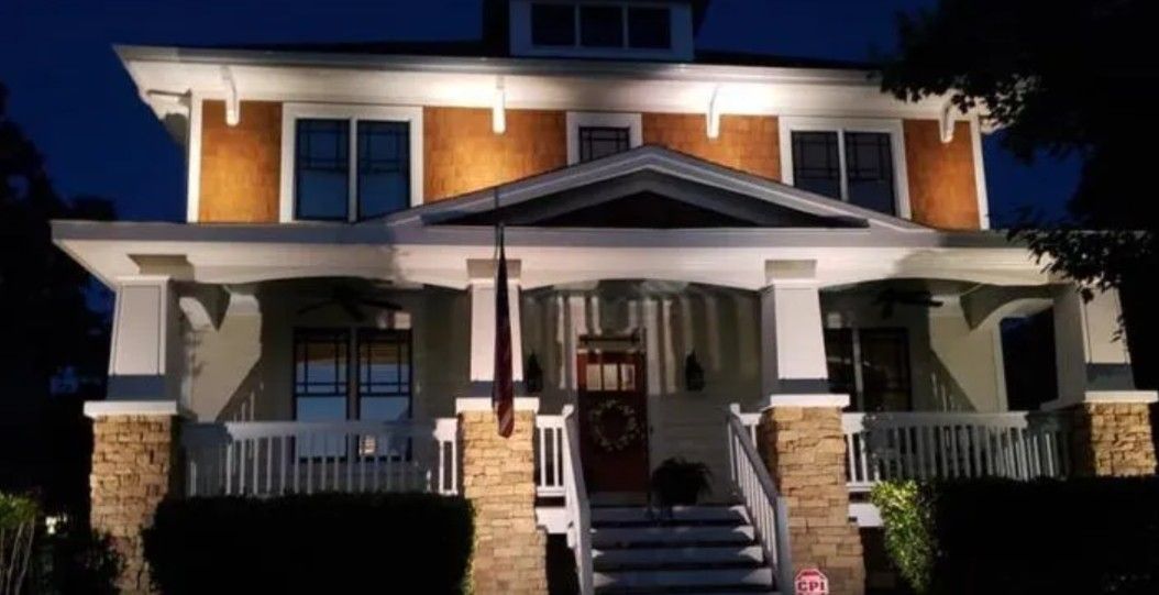 Lit-up two-story house at dusk. Beige stucco and brown brick facade, white trim, porch with stairs. Dark blue sky.