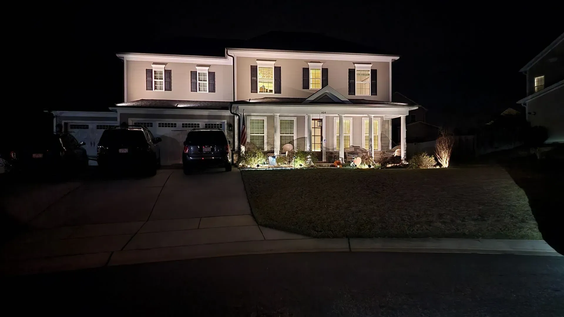 Two-story beige house at night, lit by exterior lights.  Cars in driveway.