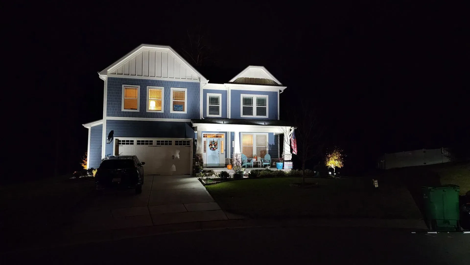 Two-story blue house at night, lit by exterior lights.  Garage on the left, porch on the right, with a black car in the driveway.