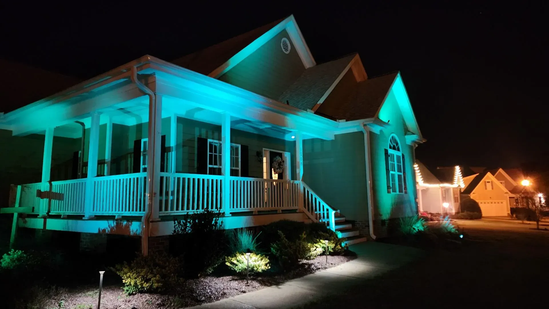 House at night illuminated with teal and green lights, porch and walkway visible.