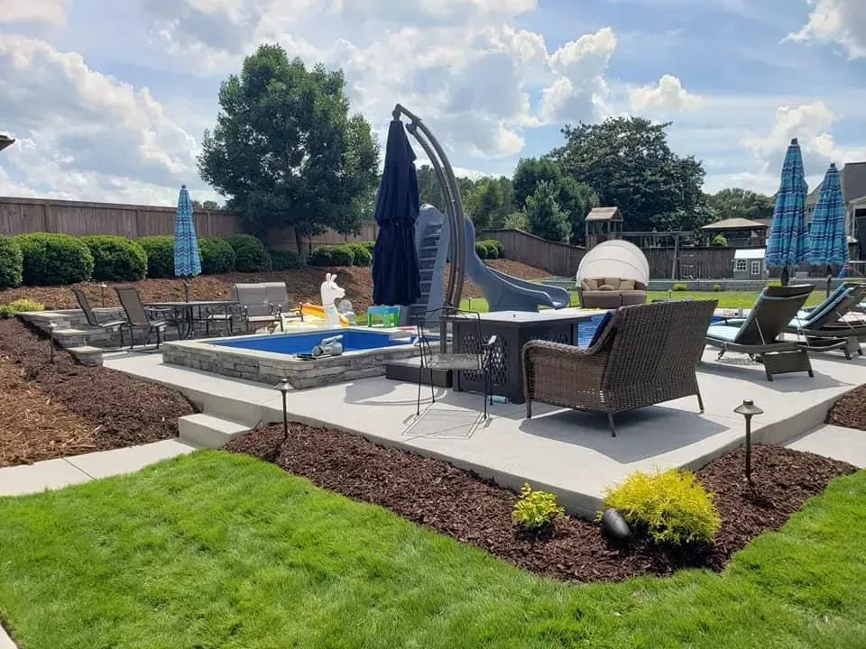 Backyard patio with hot tub, seating, and lounge chairs. Sunny day, blue umbrellas, and lush green grass.